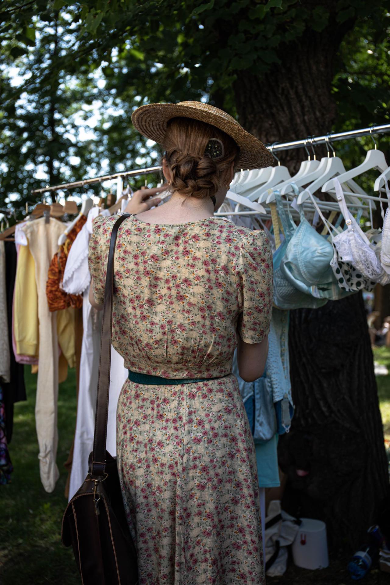 A woman in a floral vintage dress and straw hat looking through an outdoor clothes rack under a tree
